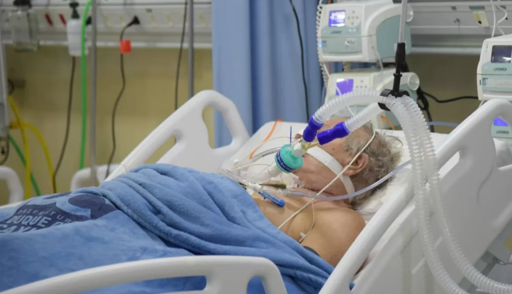 La imagen muestra a un paciente hospitalizado en una cama de cuidados intensivos, conectado a un ventilador mecánico.